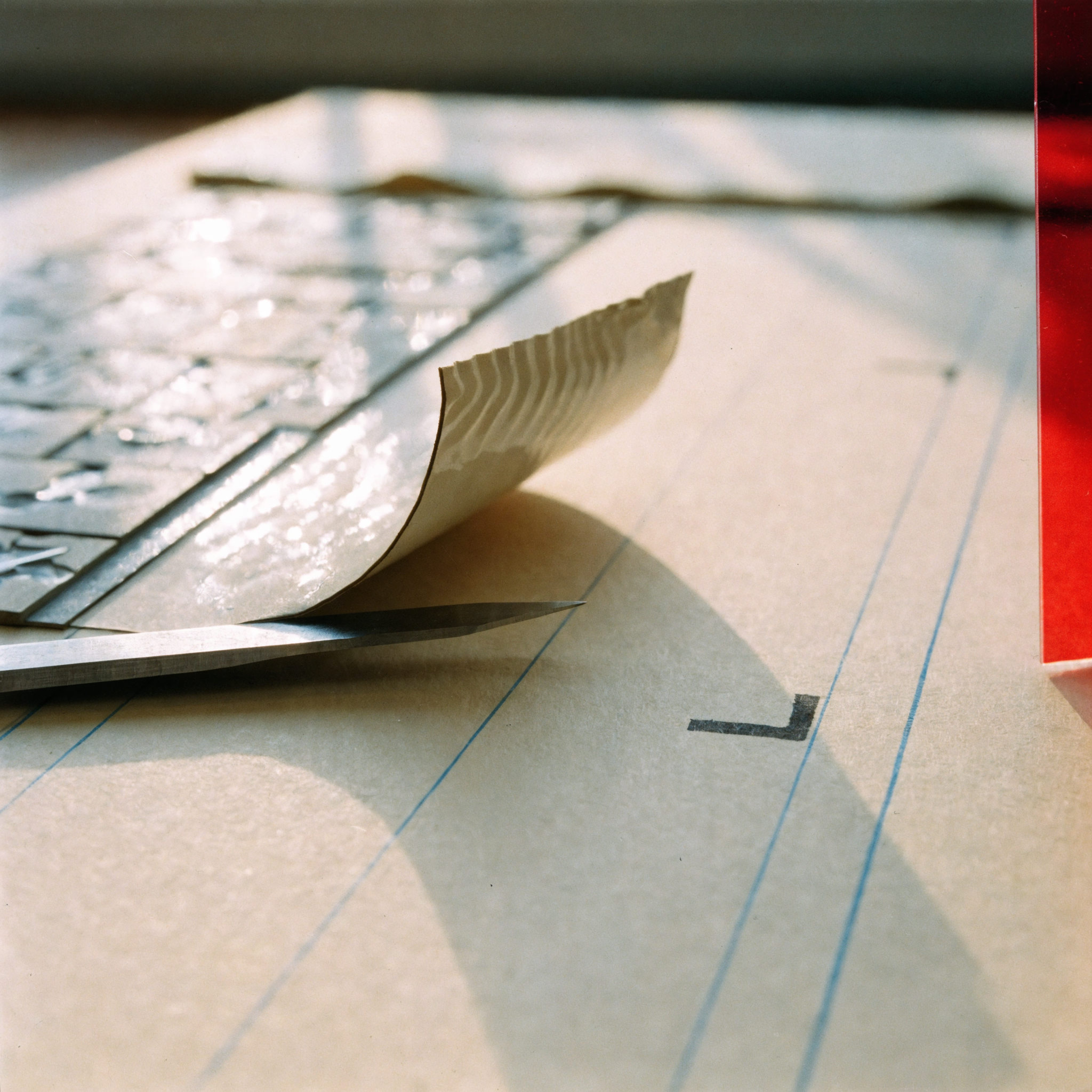 Close photograph of a knife blade lifting the curled corner of a waxed galley strip beside an inked crop mark on a cream board with light blue guides.