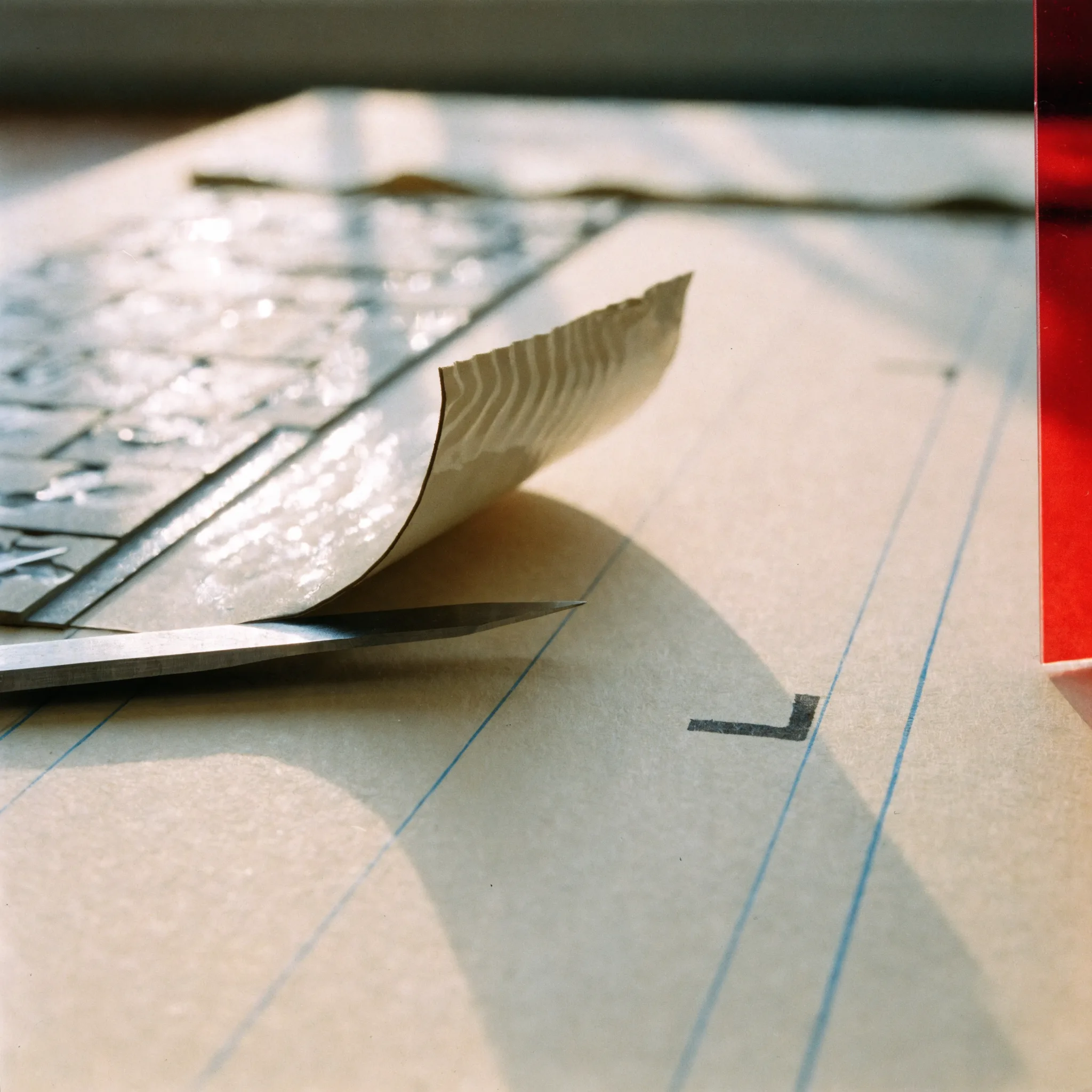 Close photograph of a knife blade lifting the curled corner of a waxed galley strip beside an inked crop mark on a cream board with light blue guides.