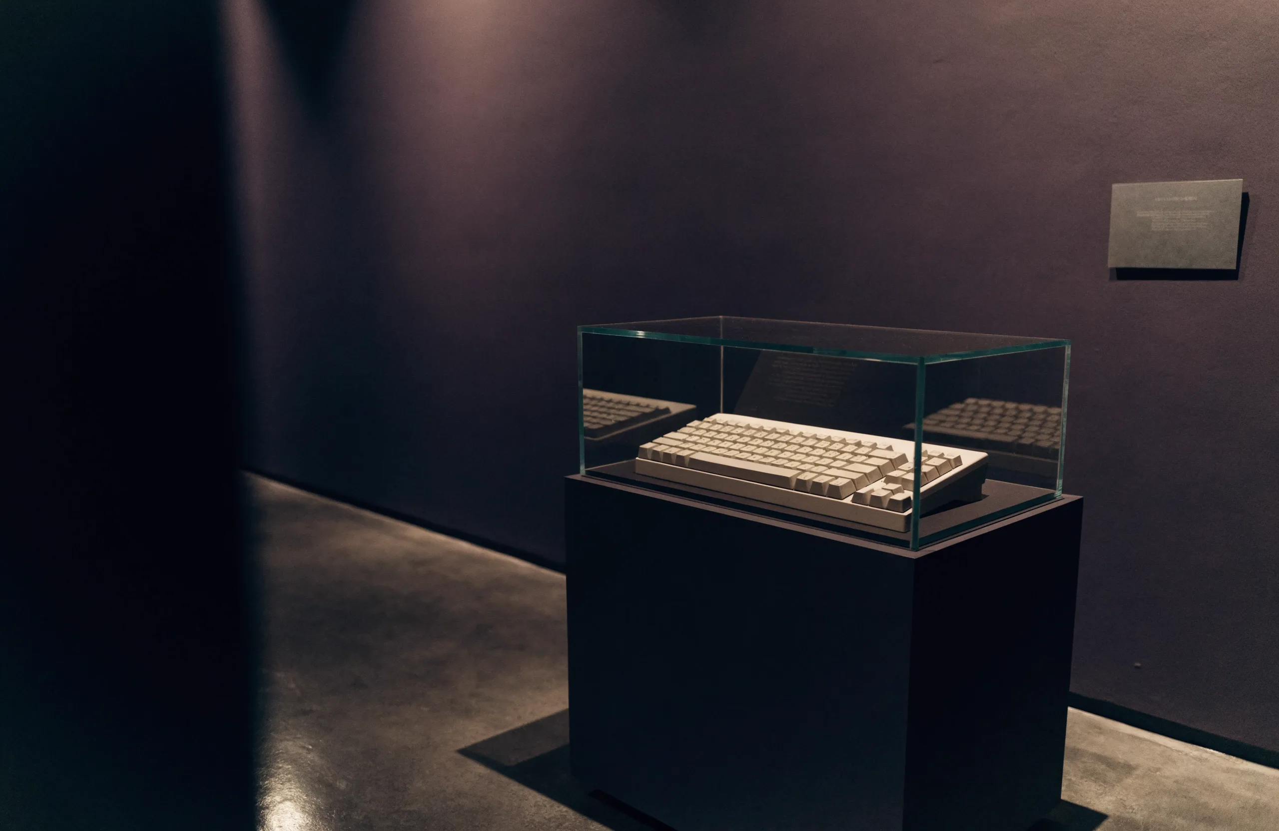 A museum vitrine in a dim gallery: a vintage beige keyboard in a glass case on a charcoal plinth, lit by a single warm spotlight.
