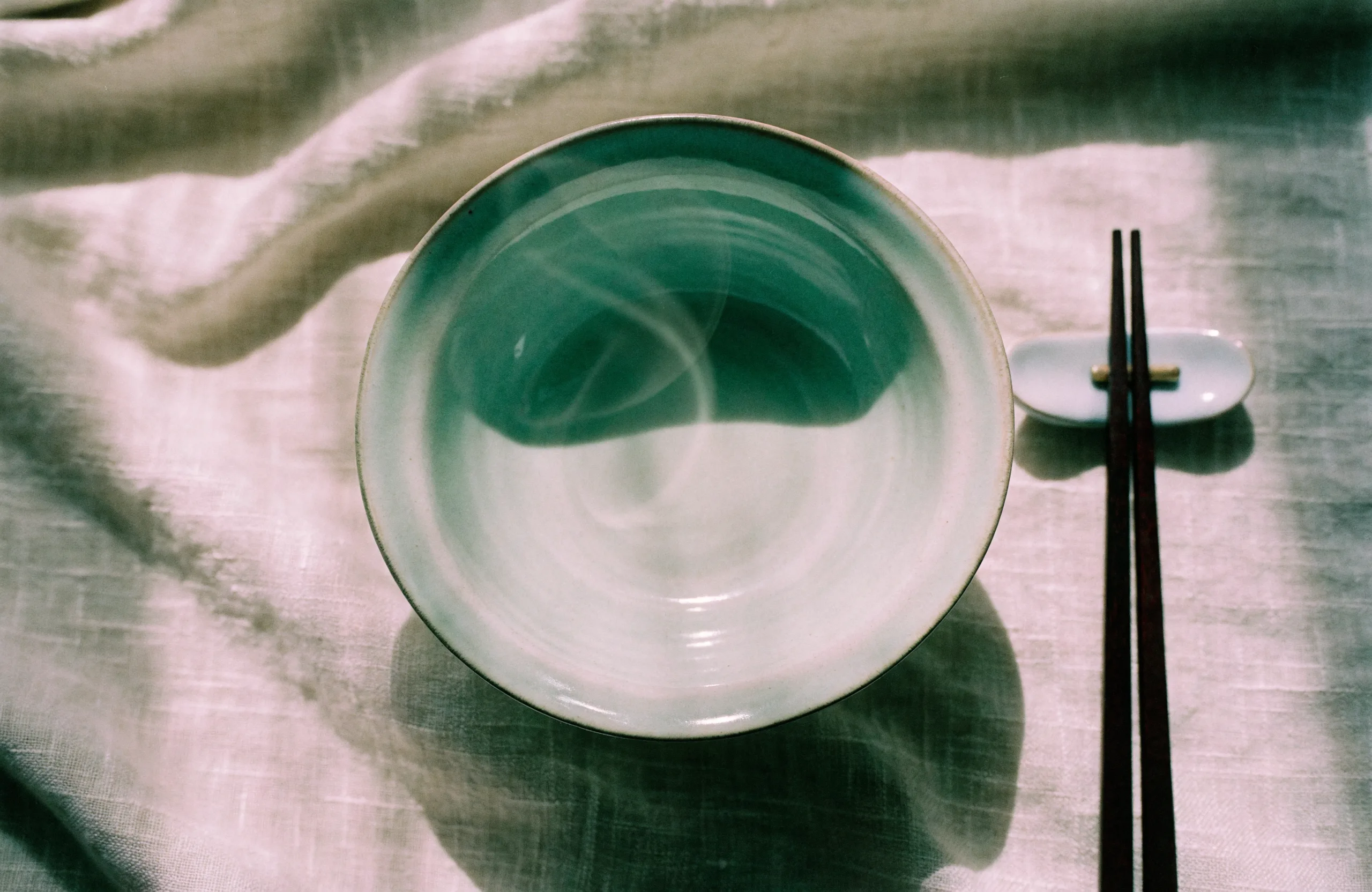 Overhead photograph of an empty celadon-glazed porcelain bowl on pale oatmeal linen, dark lacquered chopsticks resting beside it.