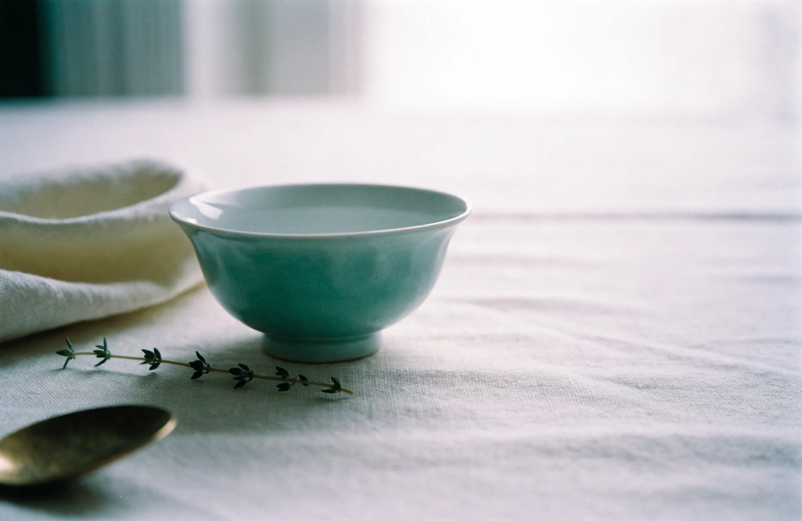 A celadon porcelain teacup beside a folded cream napkin and a sprig of herb on pale linen, soft window light.