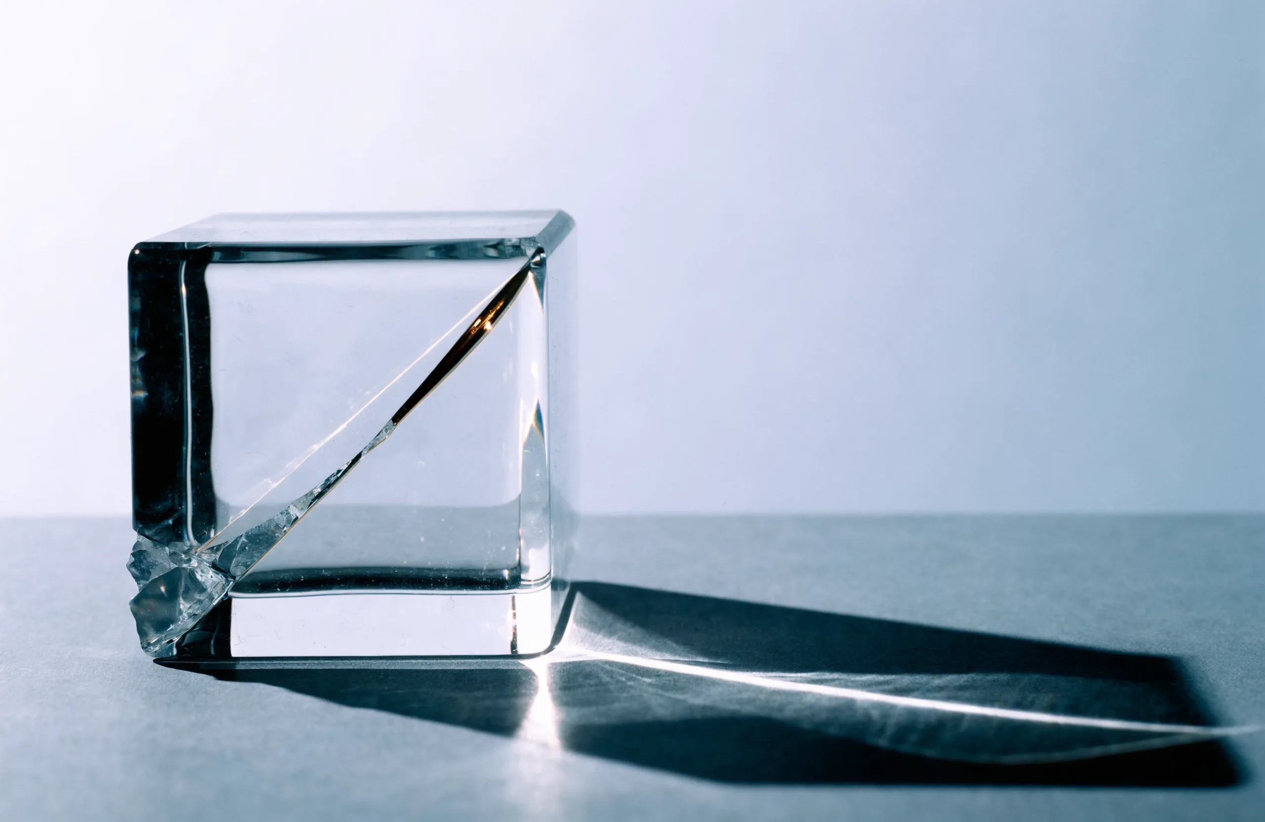 A clear glass cube with a cracked corner and a fracture line running through it, lit hard on a slate surface with a long shadow.