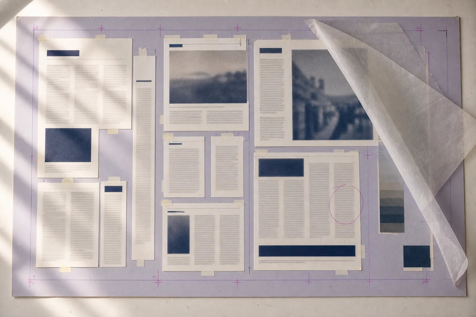 Overhead photograph of a full paste-up mechanical board with taped paper galleys, a tissue overlay folded back, and magenta crop and registration marks