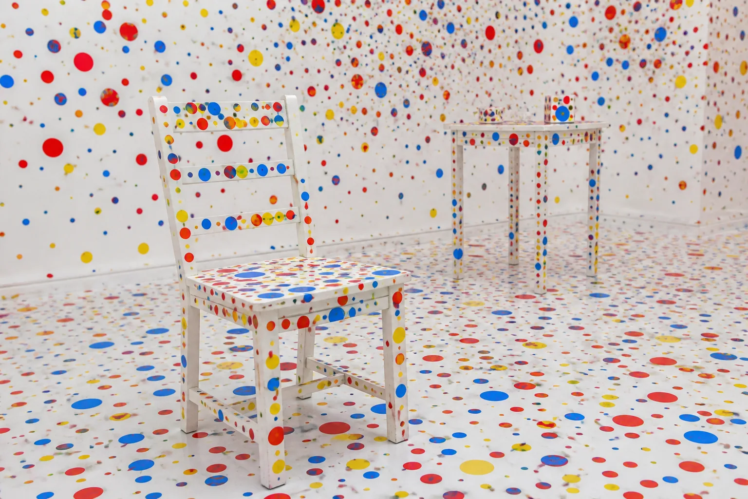 A white gallery room with a chair and table covered in red, yellow, and blue dot stickers on every surface